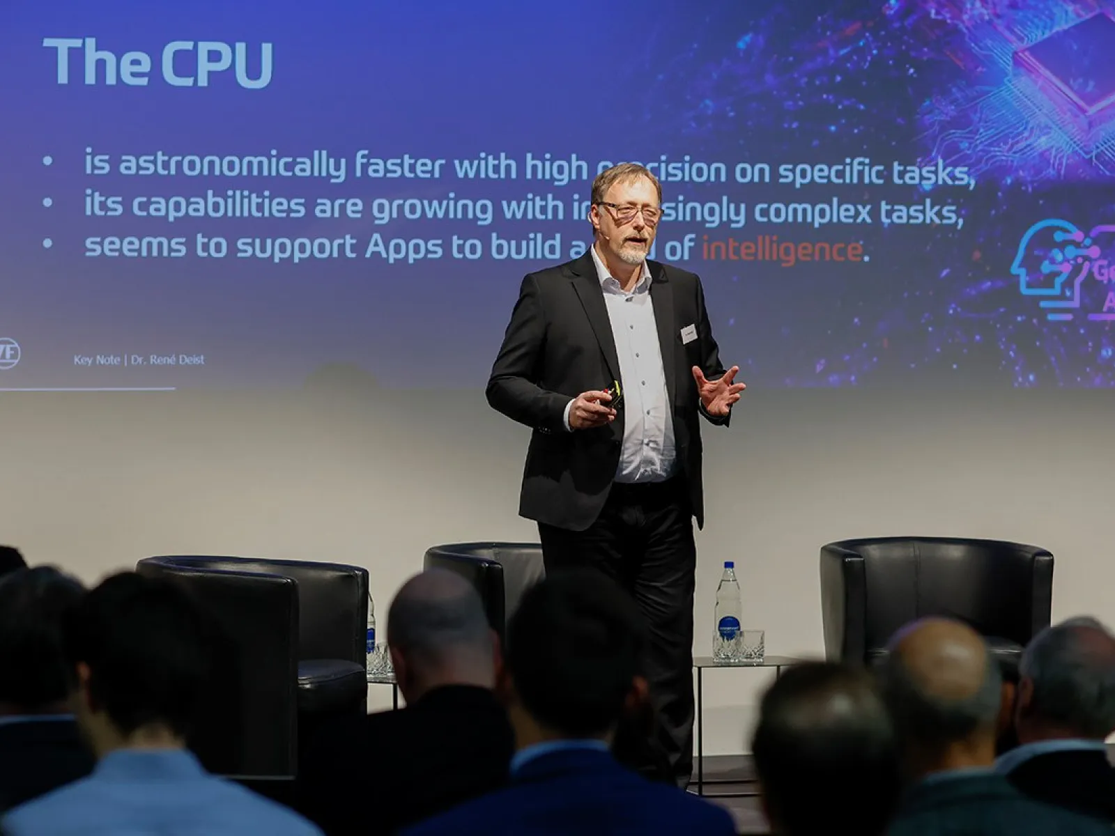 Ein Redner steht auf einer Bühne vor einem Publikum und präsentiert eine Folie über die Leistungsfähigkeit von CPUs bei einer Konferenz zur Künstlichen Intelligenz an der Universität Liechtenstein.