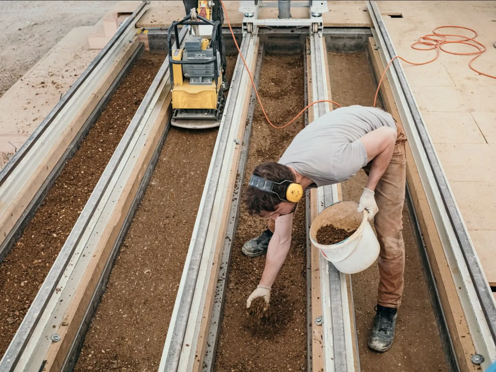 Eine Person arbeitet an einer Lehmbau-Konstruktion, indem sie Erde in eine Form füllt. Im Hintergrund ist eine Maschine zu sehen, die möglicherweise zur Verdichtung des Materials verwendet wird. Die Szene zeigt praktische Anwendung von Lehmbau-Techniken im EARTH HUB der Universität Liechtenstein.