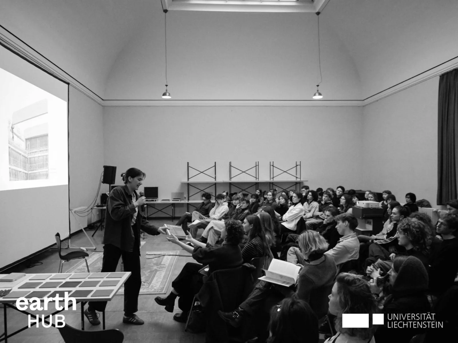 Ein Vortragender steht vor einem Publikum in einem großen Raum mit hoher Decke. Auf der Leinwand ist eine Projektion zu sehen. Im Vordergrund befindet sich ein Tisch mit Materialien. Das Logo der Universität Liechtenstein und des earth HUB ist sichtbar.