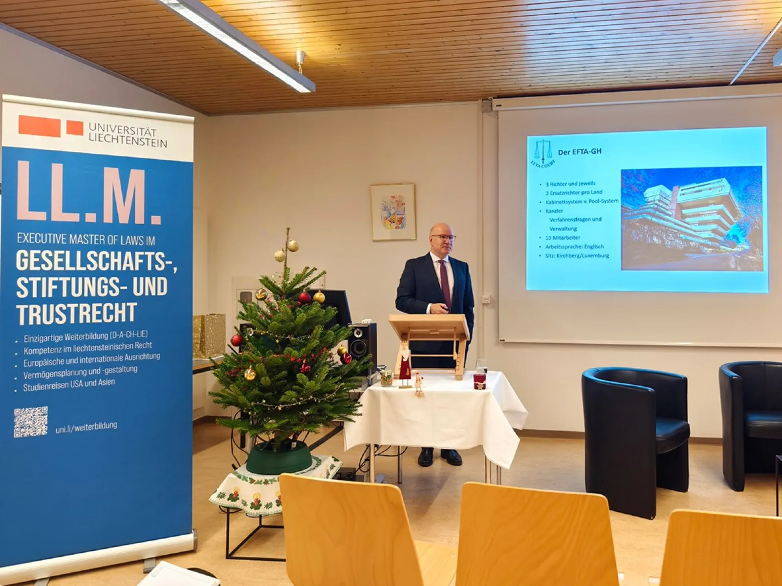 Ein Redner steht in einem Seminarraum der Universität Liechtenstein vor einem Pult. Neben ihm ist ein Banner des LL.M.-Programms für Gesellschafts-, Stiftungs- und Trustrecht zu sehen. Im Hintergrund wird eine Präsentation über den EFTA-Gerichtshof gezeigt. Ein geschmückter Weihnachtsbaum steht links im Bild.