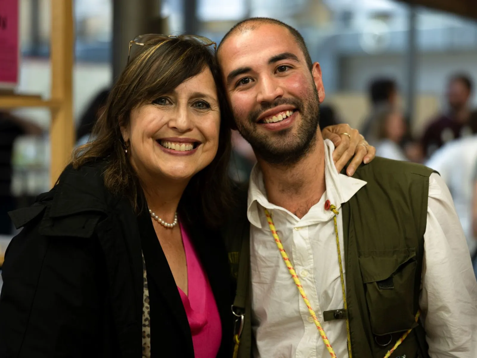 Zwei Personen lächeln in die Kamera bei einer Veranstaltung. Die Frau trägt eine Perlenkette und ein pinkes Oberteil, der Mann ein weißes Hemd. Sie stehen nah beieinander in einem Innenraum.