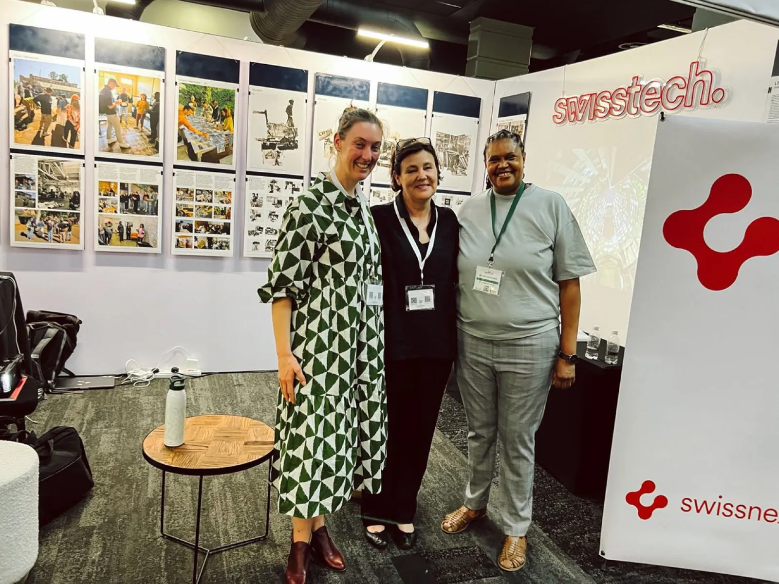 Drei Personen stehen lächelnd vor Ausstellungstafeln mit Bildern und Texten. Im Hintergrund ist ein Banner mit dem Logo von "swisstech" zu sehen. Die Szene befindet sich in einem Ausstellungsraum, der Teil der Präsentation "Making Urban Africa" auf dem South African Science Forum ist.