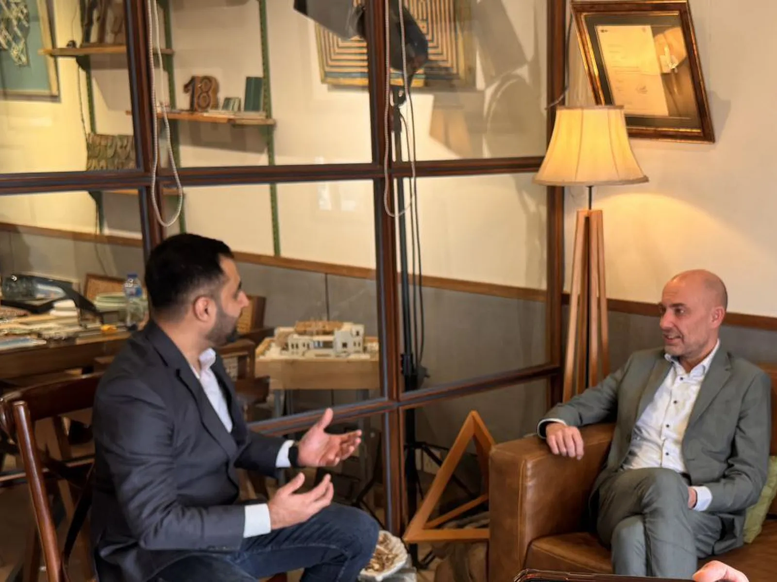 Zwei Männer in einem Gespräch in einem gemütlichen Raum mit Glaswänden und Holzmöbeln. Einer sitzt auf einem Ledersessel, der andere gestikuliert während des Gesprächs. Im Hintergrund sind Bücher und Dekorationsgegenstände zu sehen.