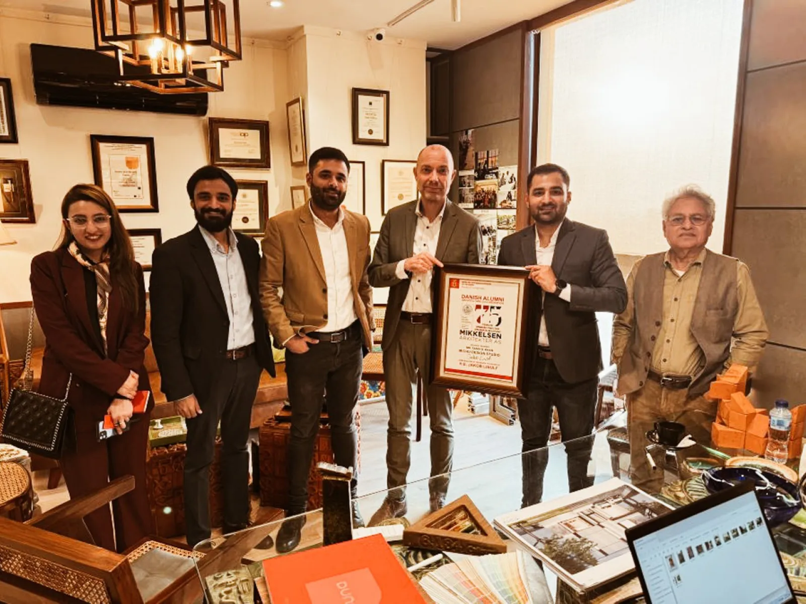 Eine Gruppe von sechs Personen steht in einem Büro, eine Person hält eine gerahmte Auszeichnung. Im Hintergrund sind Zertifikate und Auszeichnungen an der Wand zu sehen.