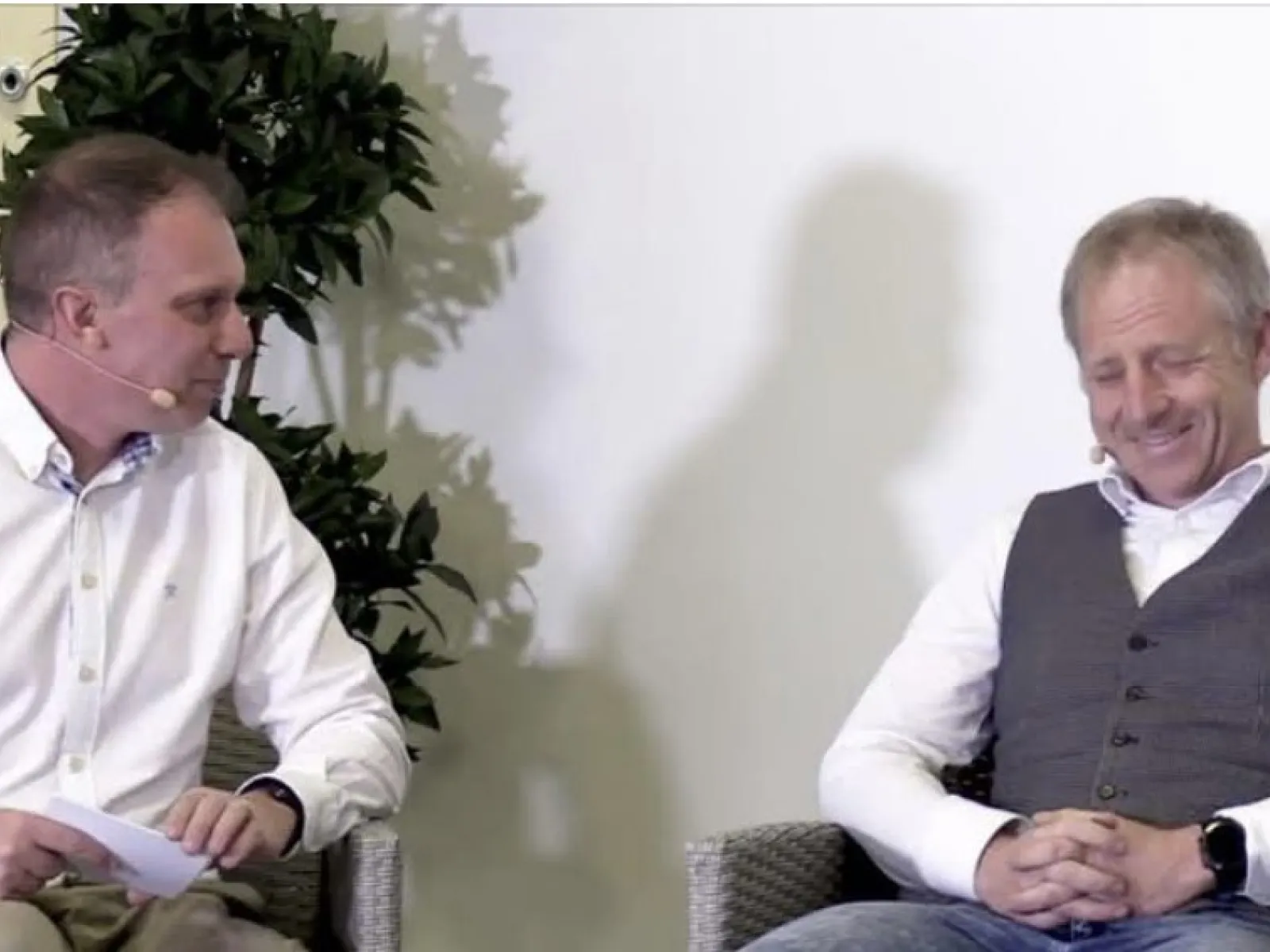 Zwei Männer sitzen auf einer Bühne und unterhalten sich. Beide tragen Mikrofone. Links sitzt ein Mann in einem weißen Hemd, rechts ein Mann in einem grauen Pullunder. Im Hintergrund ist eine Pflanze zu sehen.