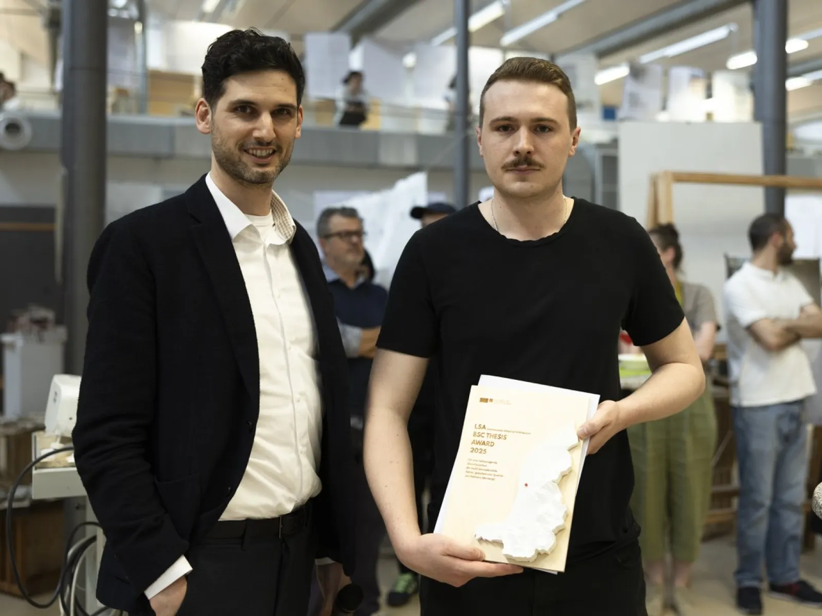 Zwei Personen stehen in einem Innenraum. Eine Person hält ein Zertifikat oder eine Auszeichnung in der Hand. Im Hintergrund sind weitere Personen und architektonische Modelle zu sehen. Die Szene wirkt wie eine Preisverleihung oder ein Event an einer Universität.