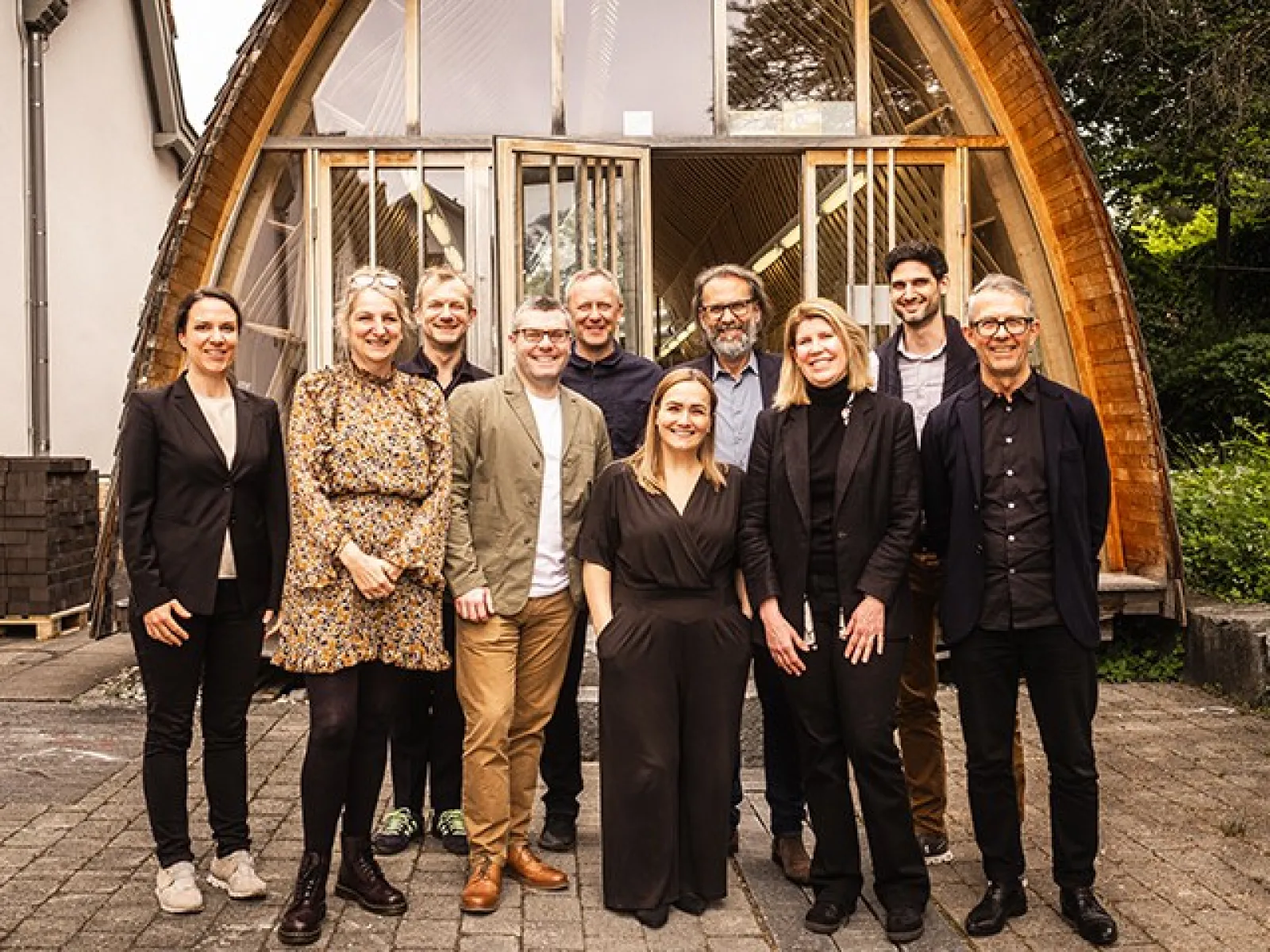 Eine Gruppe von zehn Personen steht lächelnd vor einem modernen Holzgebäude mit großen Fenstern. Die Szene vermittelt eine freundliche und professionelle Atmosphäre.