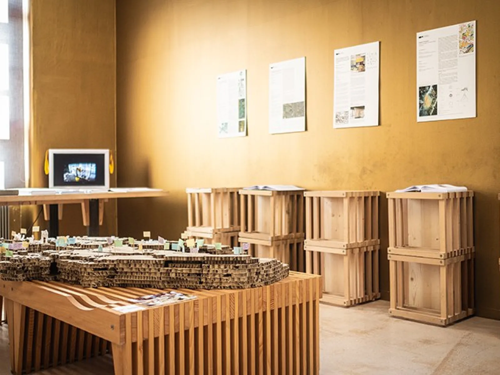 Ein Ausstellungsraum der Liechtenstein School of Architecture mit Architekturmodellen auf Holztischen und Informationsplakaten an den Wänden. Im Hintergrund ist ein Monitor zu sehen.