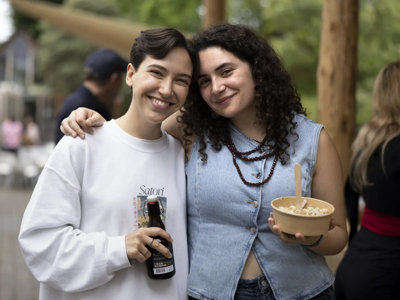 Zwei lächelnde Personen stehen eng beieinander auf einem Sommerfest. Eine Person hält eine Getränkeflasche, die andere eine Schüssel mit Essen. Im Hintergrund sind unscharfe Bäume und weitere Gäste zu sehen.