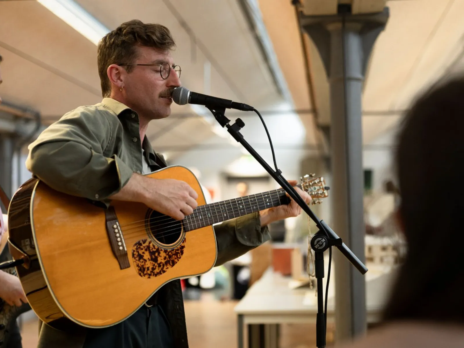 Ein Musiker spielt Gitarre und singt in ein Mikrofon bei einer Veranstaltung in einem Innenraum. Im Hintergrund sind Tische und eine helle Decke zu sehen.