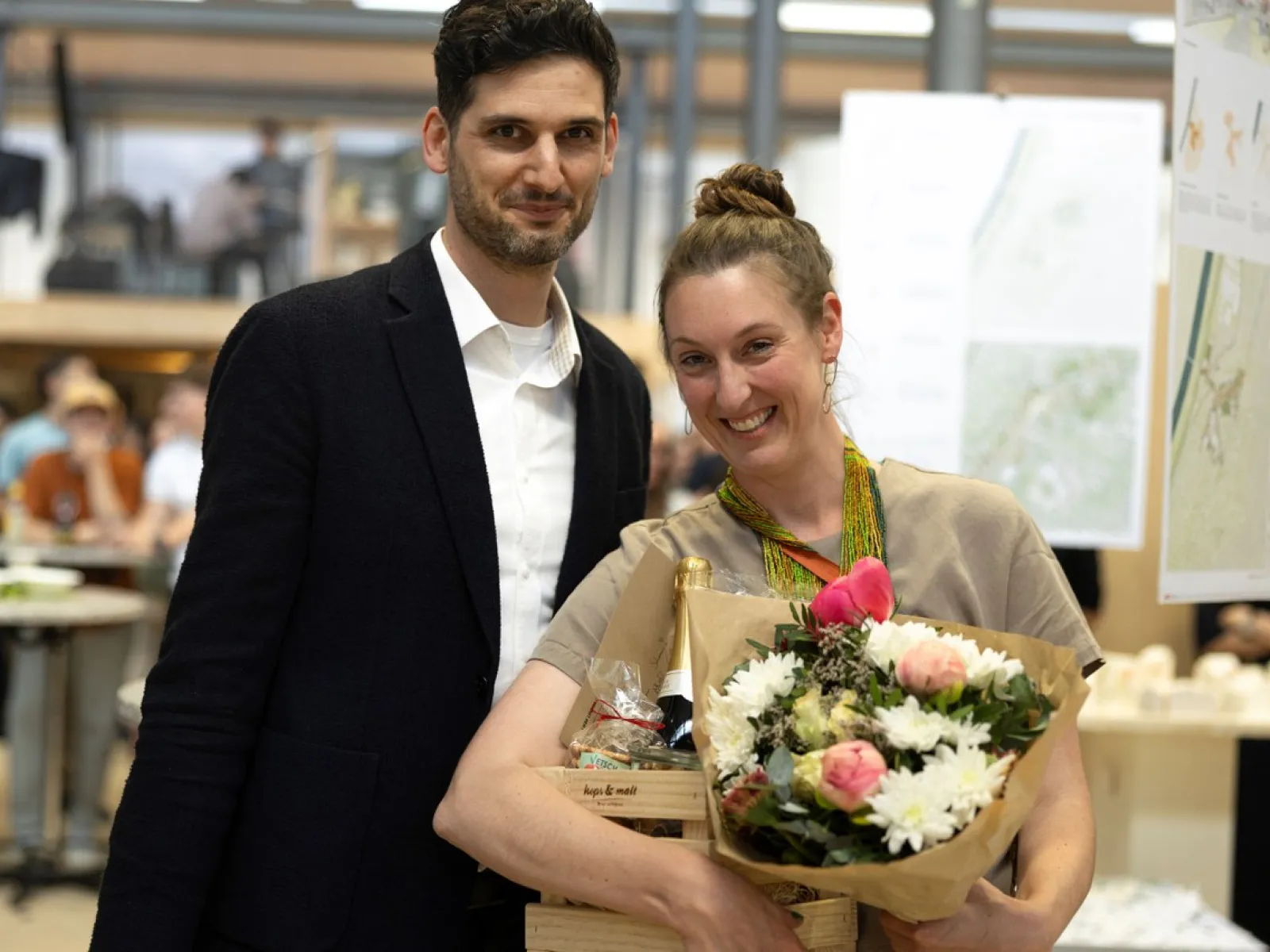 Zwei Personen stehen lächelnd in einem Innenraum. Eine Person hält einen Blumenstrauß und eine Holzkiste mit Geschenken. Im Hintergrund sind unscharf architektonische Pläne und weitere Personen zu erkennen.