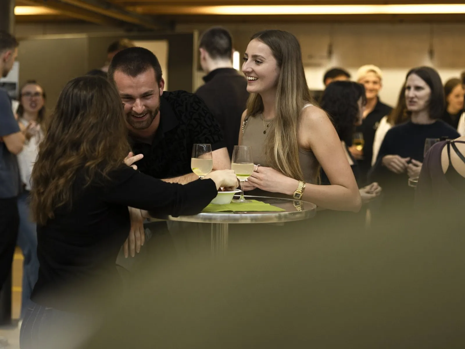 Eine Gruppe von Menschen unterhält sich fröhlich bei einem Stehempfang. Zwei Personen halten Gläser mit Getränken in der Hand. Im Hintergrund sind weitere Gäste zu sehen. Die Stimmung wirkt entspannt und gesellig.
