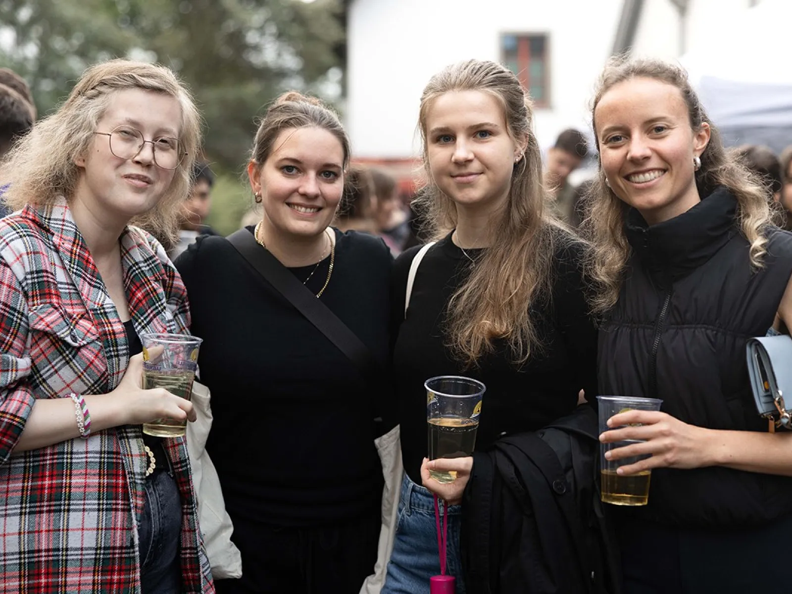 Vier junge Frauen stehen lächelnd zusammen auf einem Sommerfest der Universität Liechtenstein. Sie halten Getränke in der Hand und sind von anderen Feiernden umgeben. Im Hintergrund sind Bäume und ein Gebäude zu sehen.
