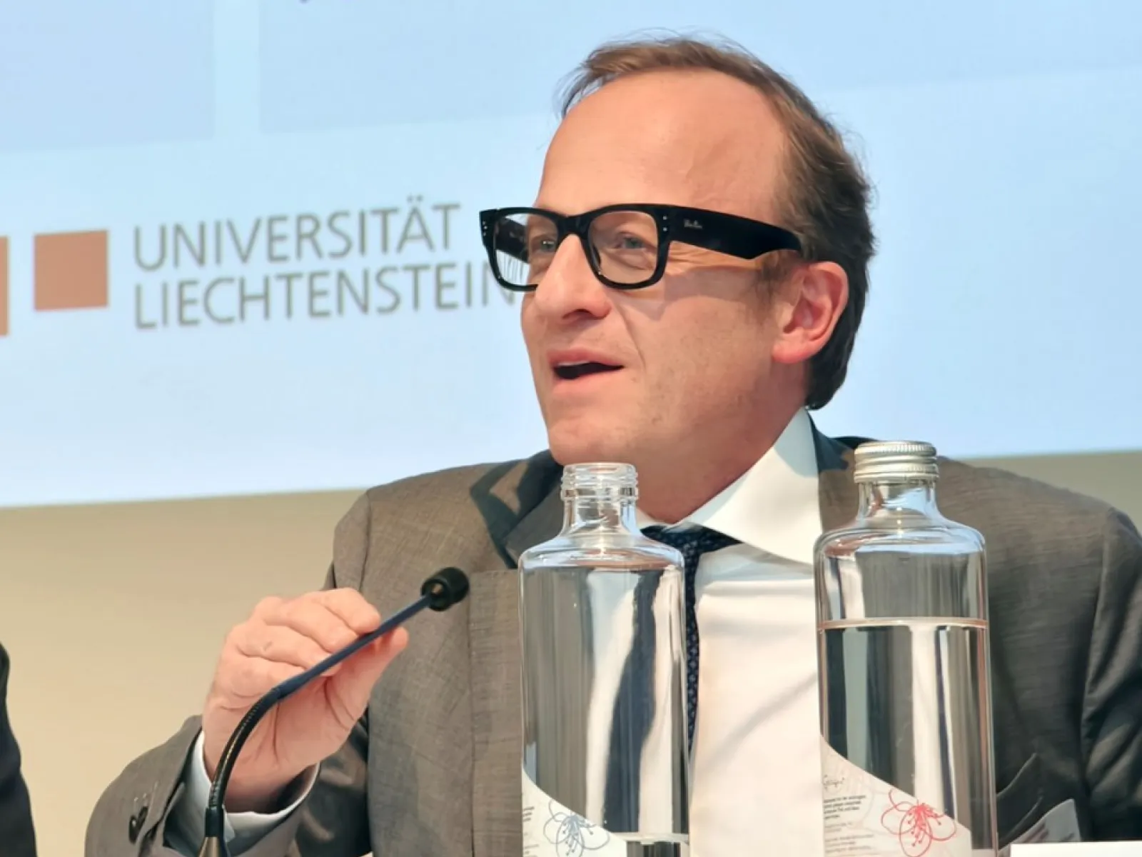 Ein Mann in Anzug und Brille spricht in ein Mikrofon auf einer Konferenz im Auditorium der Universität Liechtenstein. Vor ihm stehen Wasserflaschen. Im Hintergrund ist das Logo der Universität zu sehen.