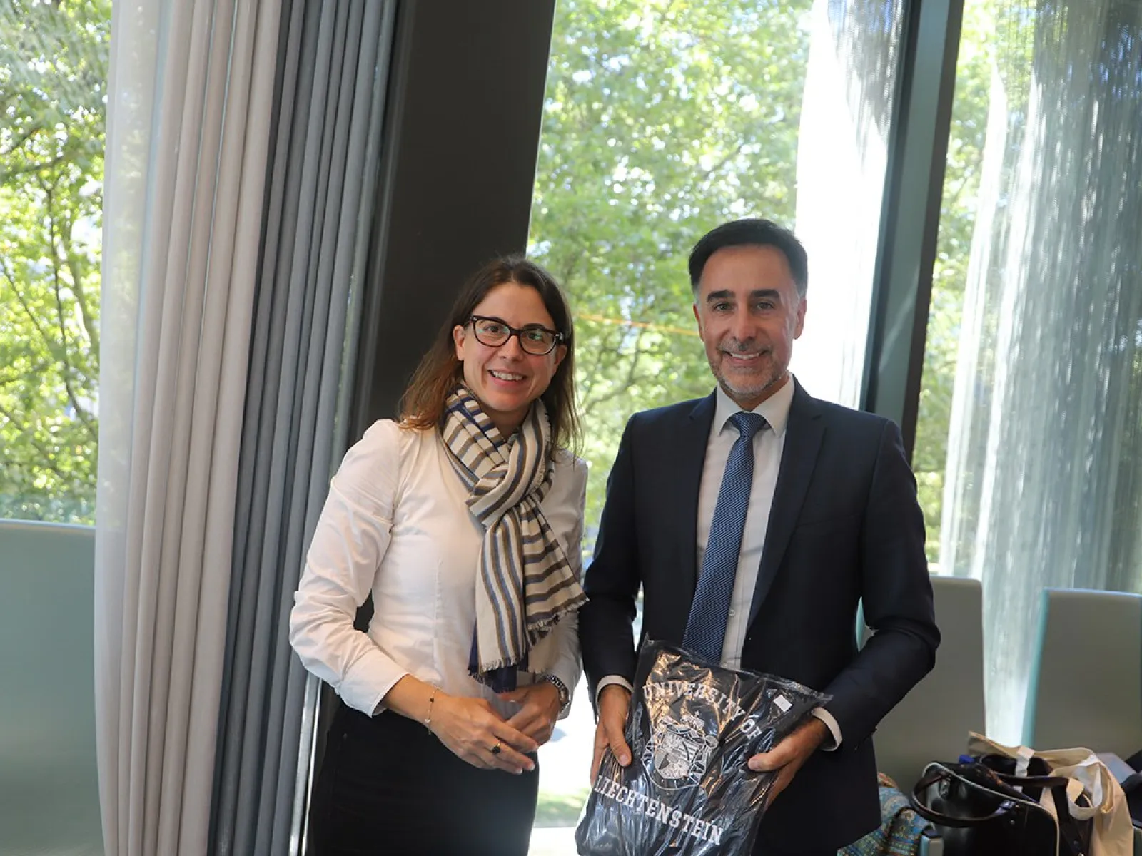 Zwei Personen stehen lächelnd in einem modernen Raum mit großen Fenstern. Eine Person hält eine Tasche mit dem Logo der Universität Liechtenstein. Im Hintergrund sind Bäume durch die Fenster sichtbar.