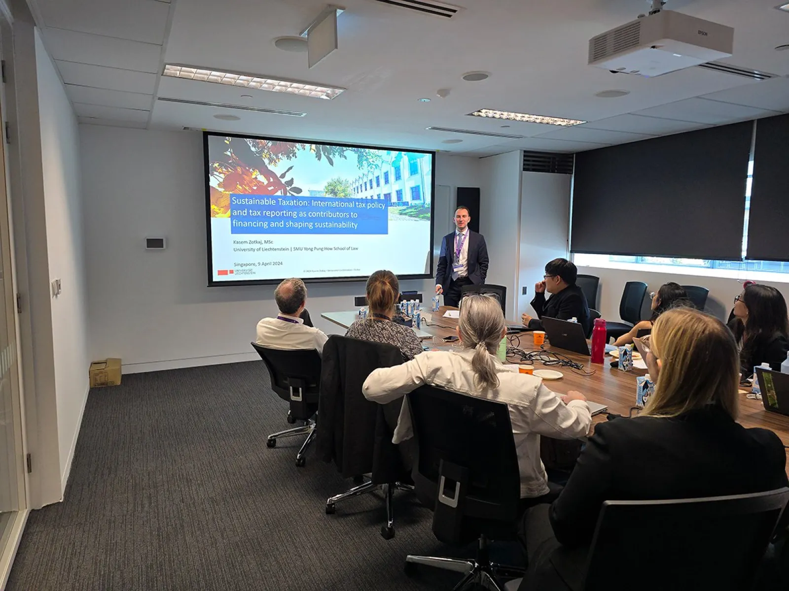 Ein Vortragender steht vor einer Präsentation mit dem Titel "Sustainable Taxation" in einem Konferenzraum. Mehrere Personen sitzen an einem Tisch und hören zu. Die Präsentation behandelt internationale Steuerpolitik und Nachhaltigkeit.