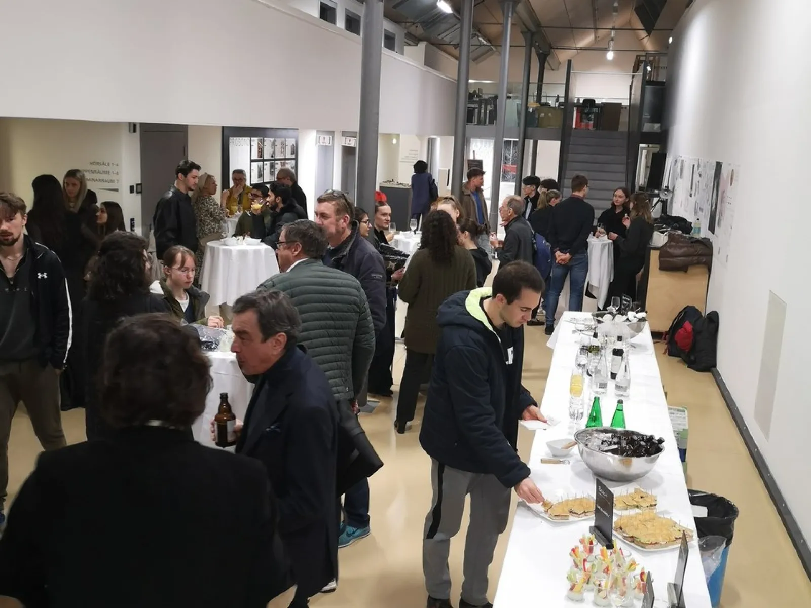 Eine Gruppe von Menschen steht in einem modernen Veranstaltungsraum, unterhält sich und genießt ein Buffet mit Getränken und Snacks. Im Hintergrund sind hohe Decken und eine Uhr sichtbar.