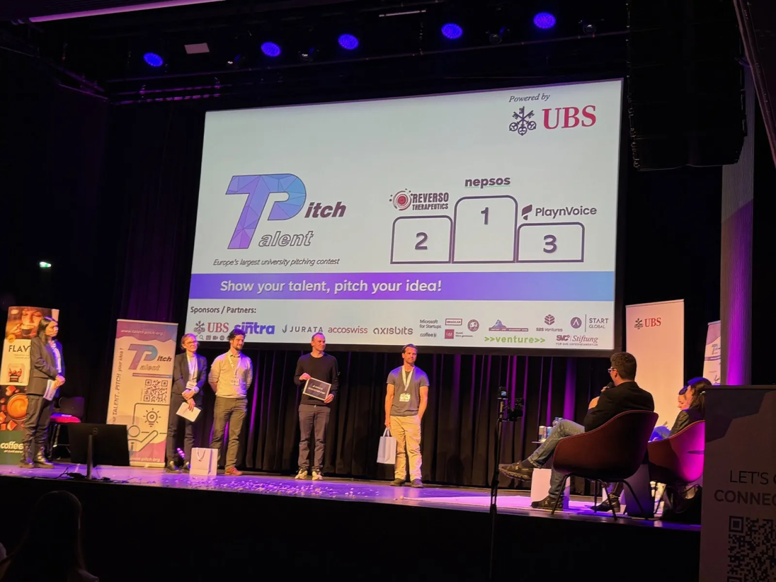 Eine Bühne mit vier Personen, die an einem Pitch-Wettbewerb teilnehmen. Im Hintergrund ist eine Leinwand mit dem Logo von "Talent Pitch" und den Namen der Start-ups "Reverso Therapeutics", "nepsos" und "PlaynVoice" zu sehen. Das Event wird von UBS gesponsert. Zuschauer sitzen auf der rechten Seite.