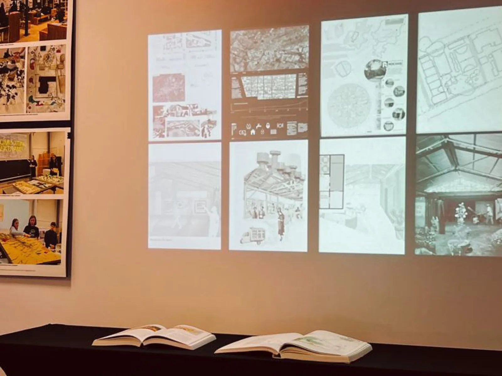 Ein Ausstellungstisch mit offenen Büchern steht vor einer Wand, auf der Architekturzeichnungen und Pläne projiziert sind. Links sind Fotos von Designprojekten zu sehen. Oben leuchtet ein "swisstech" Schild in roter Schrift.