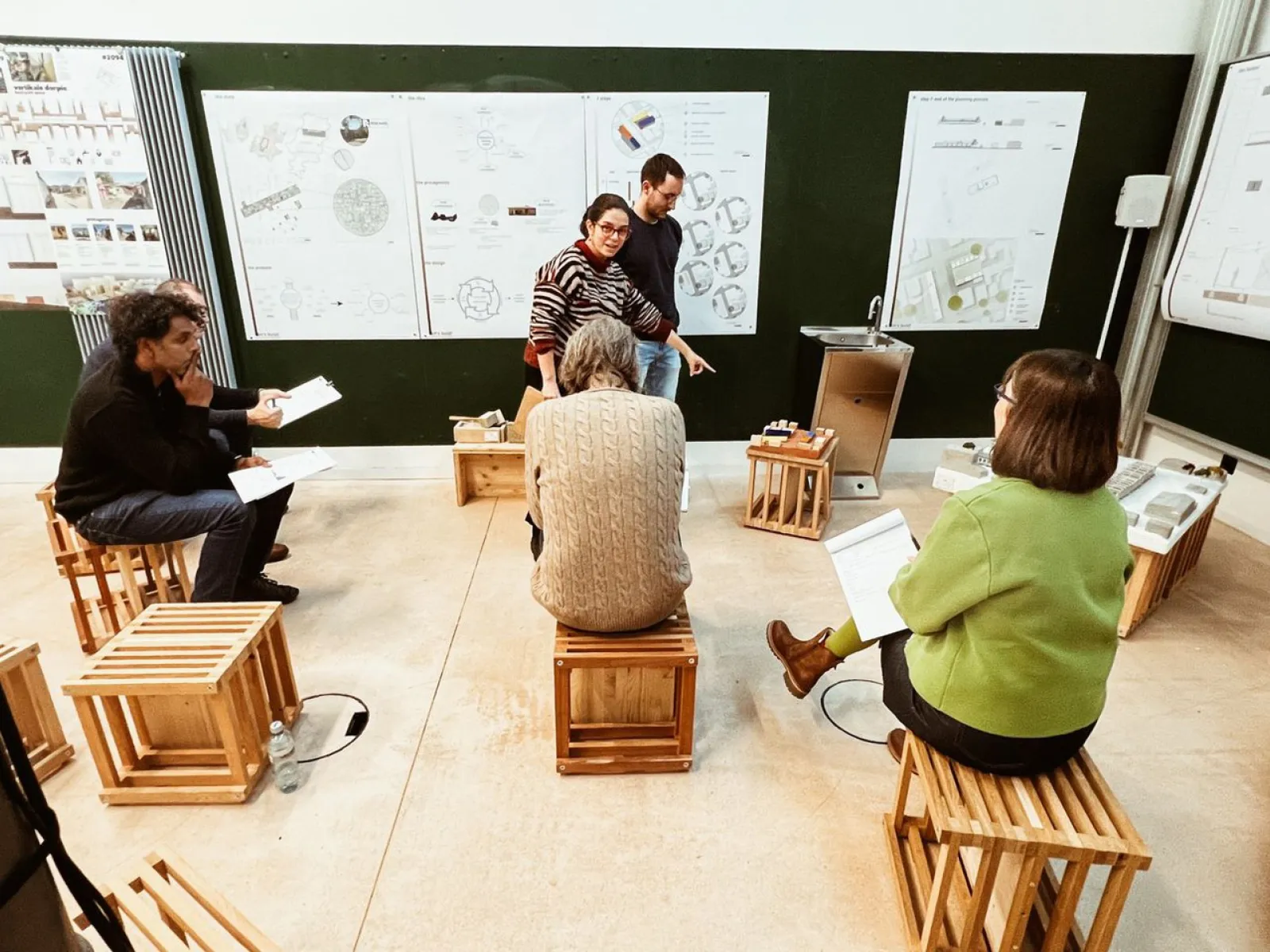Eine Gruppe von sechs Personen sitzt auf Holzkisten in einem Raum mit Plakaten an der Wand. Zwei Personen stehen und diskutieren, während die anderen Notizen machen. Der Raum wirkt wie ein Workshop oder eine Präsentation.