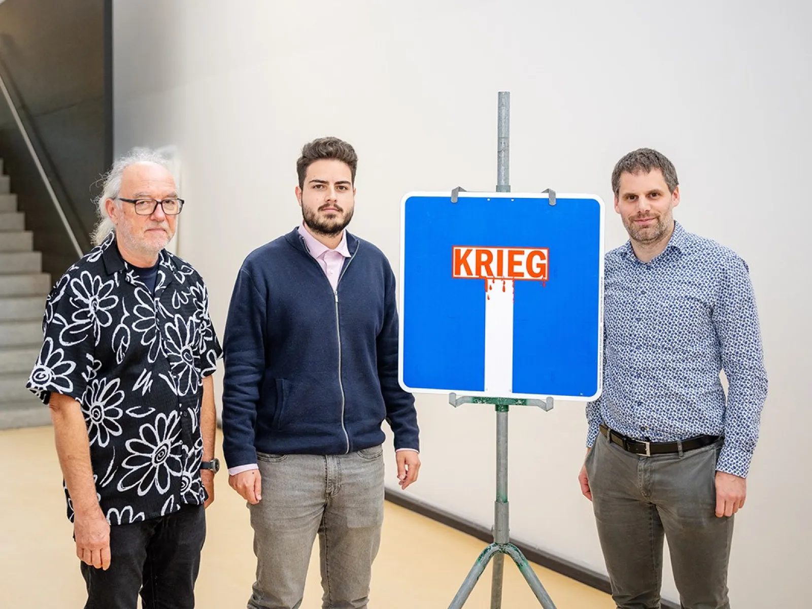 Drei Männer stehen neben einem blauen Verkehrsschild mit der Aufschrift "Krieg", das symbolisch eine Sackgasse darstellt. Das Bild wurde bei der Veranstaltung "Verkehrsschilder der Gerechtigkeit" an der Universität Liechtenstein aufgenommen.