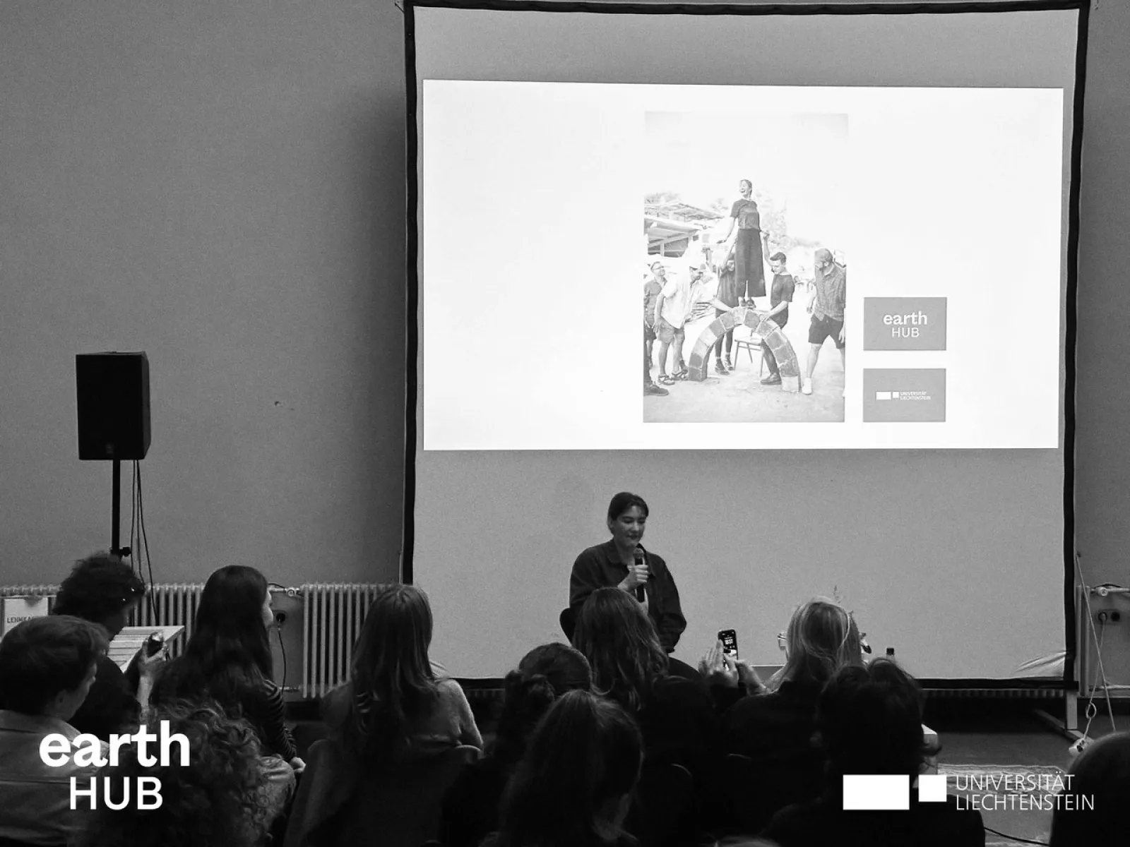 Eine Vortragende steht vor einem Publikum und präsentiert. Im Hintergrund ist eine Leinwand zu sehen. Das Bild ist in schwarz-weiss.