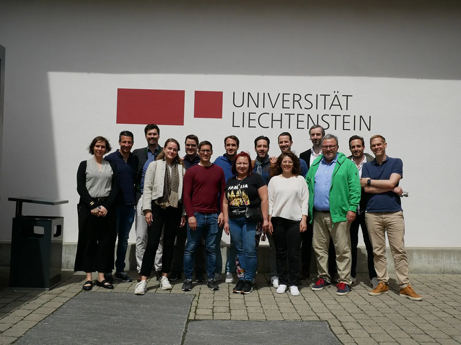Gruppenfoto von Alumni des Abschlussjahrgangs 2013 des Masters in Banking and Financial Management vor dem Logo der Universität Liechtenstein. Die Personen stehen lächelnd zusammen auf dem Campus bei sonnigem Wetter.