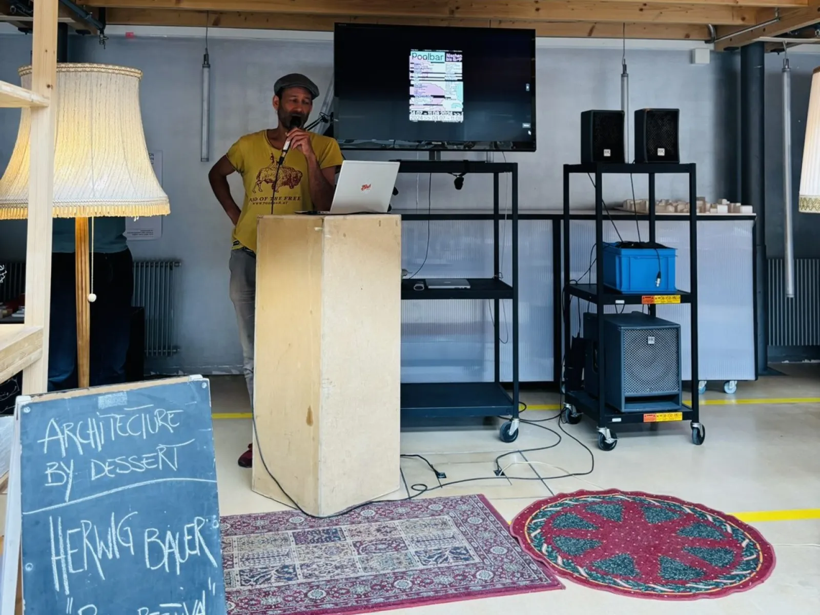 Ein Mann steht an einem Rednerpult in einem Raum mit Holzdach. Neben ihm ist ein Bildschirm mit einer Präsentation und ein Schild mit der Aufschrift „Architecture by Dessert, Herwig Bauer, Poolbarfestival“. Der Raum ist mit Teppichen und Stehlampen dekoriert.