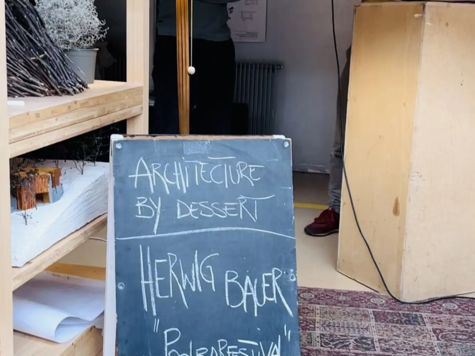 Ein Mann spricht in ein Mikrofon neben einem Rednerpult. Im Vordergrund steht eine Tafel mit der Aufschrift „Architecture by Dessert, Herwig Bauer, Poolbarfestival“. Links sind ein Regal und eine Stehlampe zu sehen.