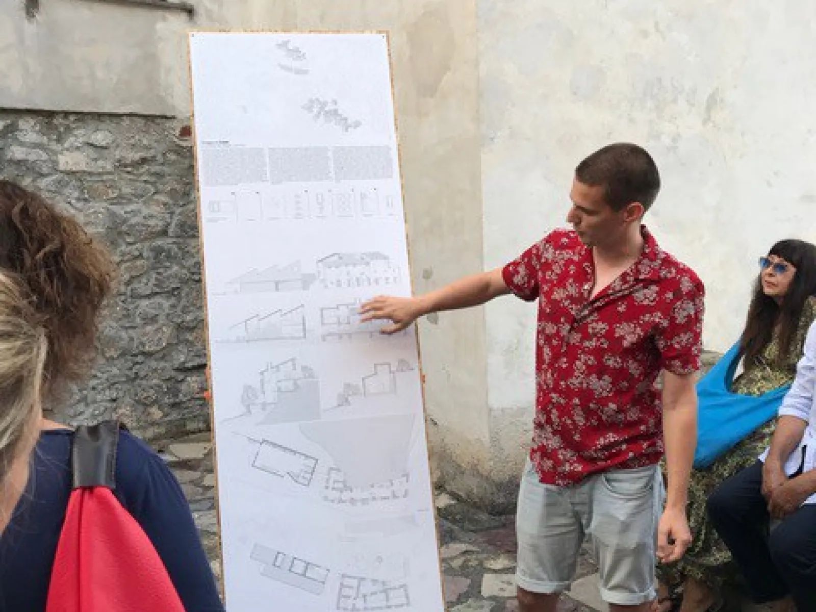 Ein Student präsentiert architektonische Skizzen auf einem großen Plakat in einem historischen Dorf. Um ihn herum stehen und sitzen mehrere Personen, die aufmerksam zuhören. Die Szene spielt sich in einem gepflasterten Außenbereich ab.