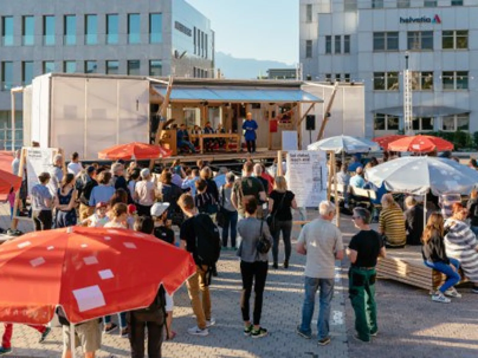 Eine Menschenmenge versammelt sich vor einer modularen Plattform im Freien, umgeben von modernen Gebäuden. Die Plattform dient als Veranstaltungsort mit Rednern und Sitzgelegenheiten. Bunte Sonnenschirme bieten Schatten. Die Szene zeigt eine lebendige, urbane Umgebung, die für Austausch und Beteiligung genutzt wird.