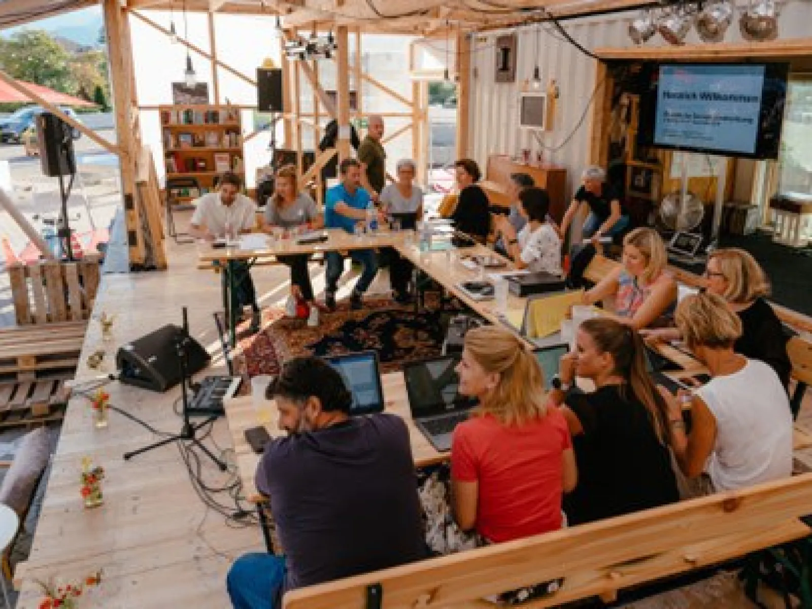 Eine Gruppe von Menschen sitzt in einem offenen, modularen Raum an einem Tisch mit Laptops. Der Raum ist aus Holz und enthält Bücherregale und technische Ausrüstung. Es herrscht eine lebhafte Diskussionsatmosphäre.