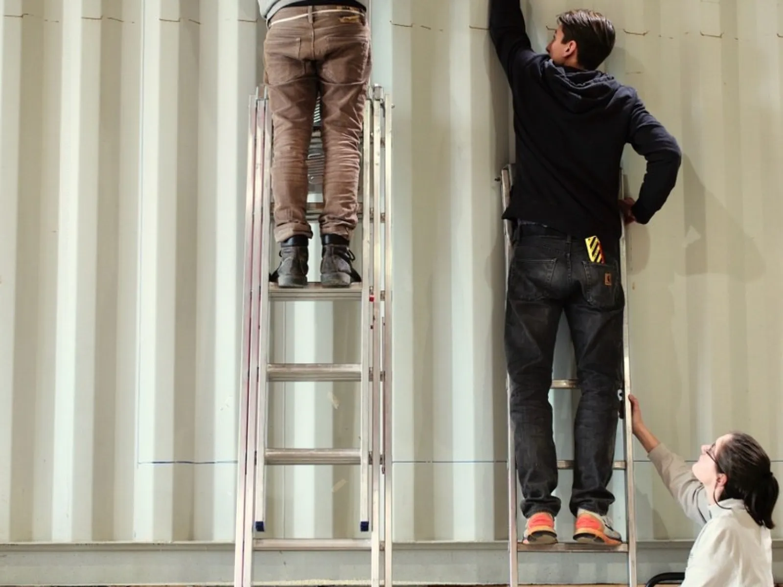 Drei Personen arbeiten an einem großen, weißen Container. Zwei stehen auf Leitern und befestigen etwas an der Wand, während die dritte Person unten steht und assistiert.