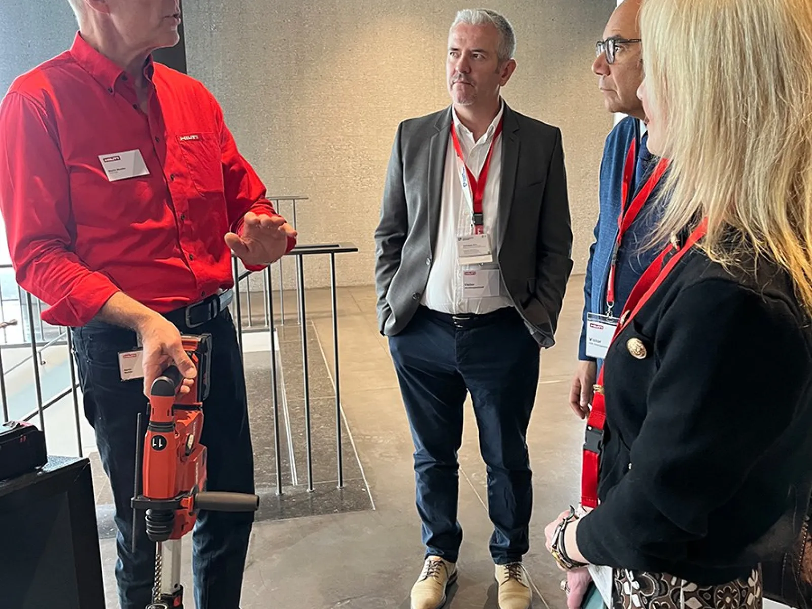 Eine Gruppe von vier Personen steht in einem modernen Gebäude. Eine Person hält ein Werkzeug und erklärt etwas den anderen. Alle tragen Namensschilder und hören aufmerksam zu.