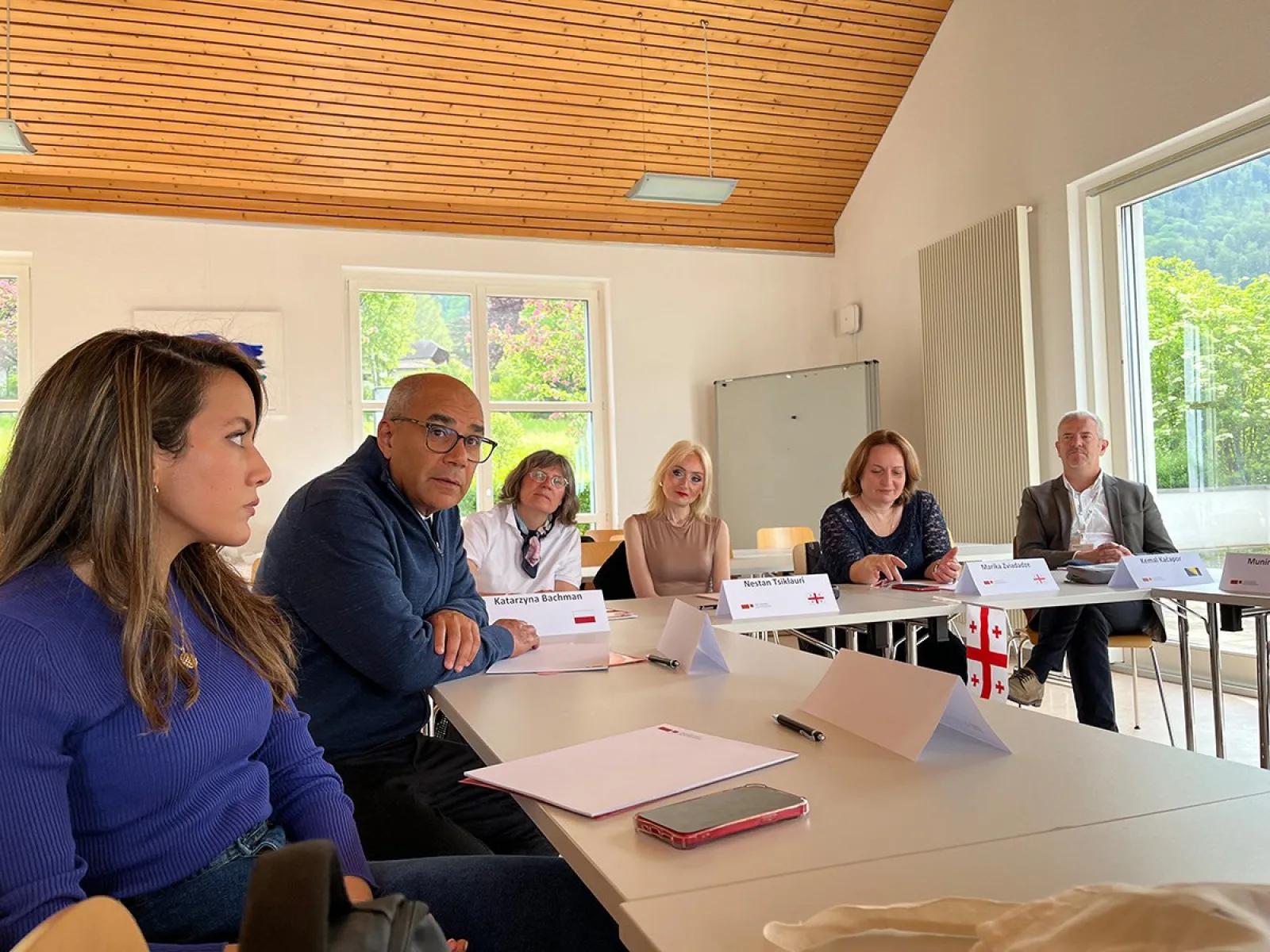 Eine Gruppe von Personen sitzt an einem Konferenztisch in einem hellen Raum mit großen Fenstern. Sie diskutieren angeregt, vor ihnen liegen Notizblöcke und Namensschilder. Im Hintergrund sind grüne Bäume und Berge zu sehen.