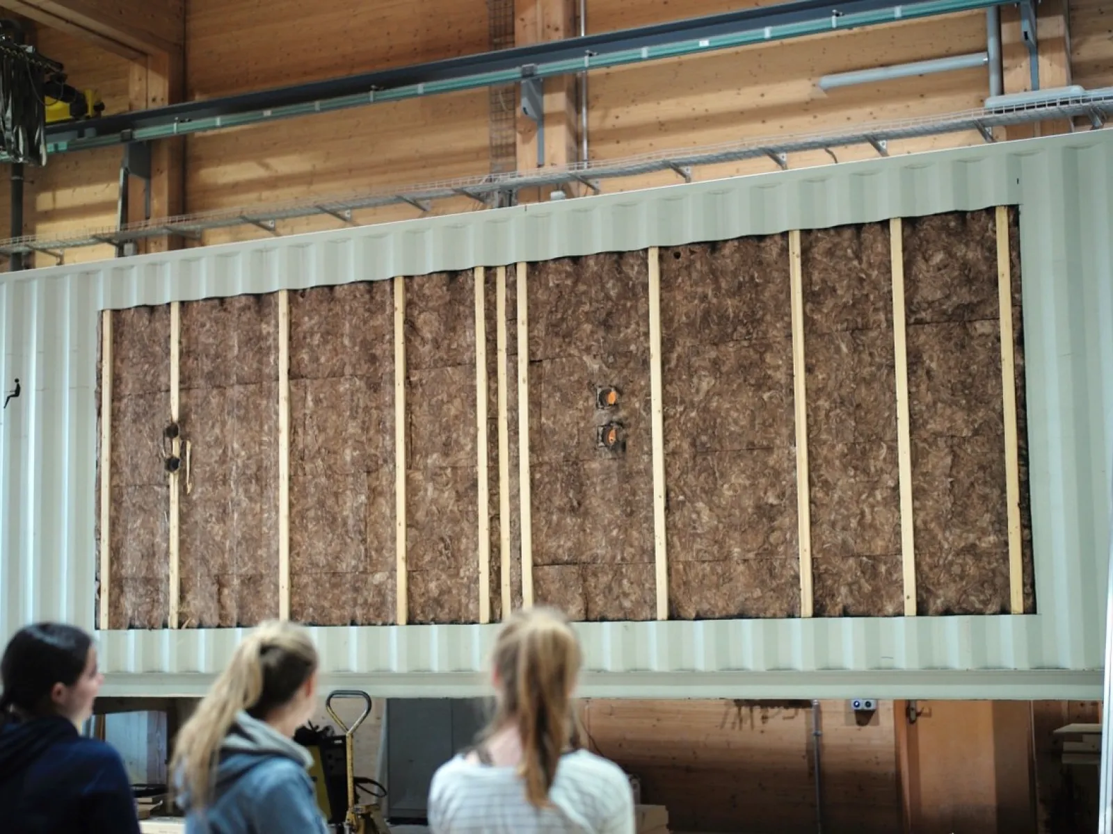Zwei Personen betrachten eine große, isolierte Wandstruktur in einem Innenraum. Die Wand besteht aus Holz und Dämmmaterial und ist Teil eines modularen Bauprojekts.