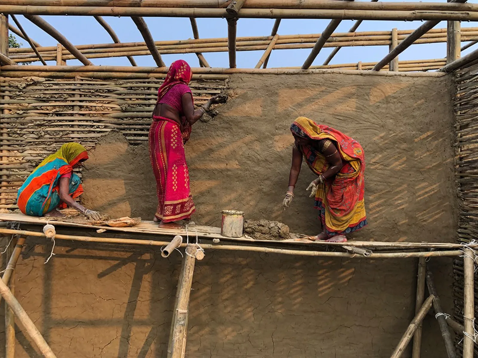 Drei Frauen in bunten Saris arbeiten auf einem Bambusgerüst an einer Lehmwand. Sie tragen Lehm auf die Wand auf, um eine Schule in Bahuarwa, Indien, zu bauen.