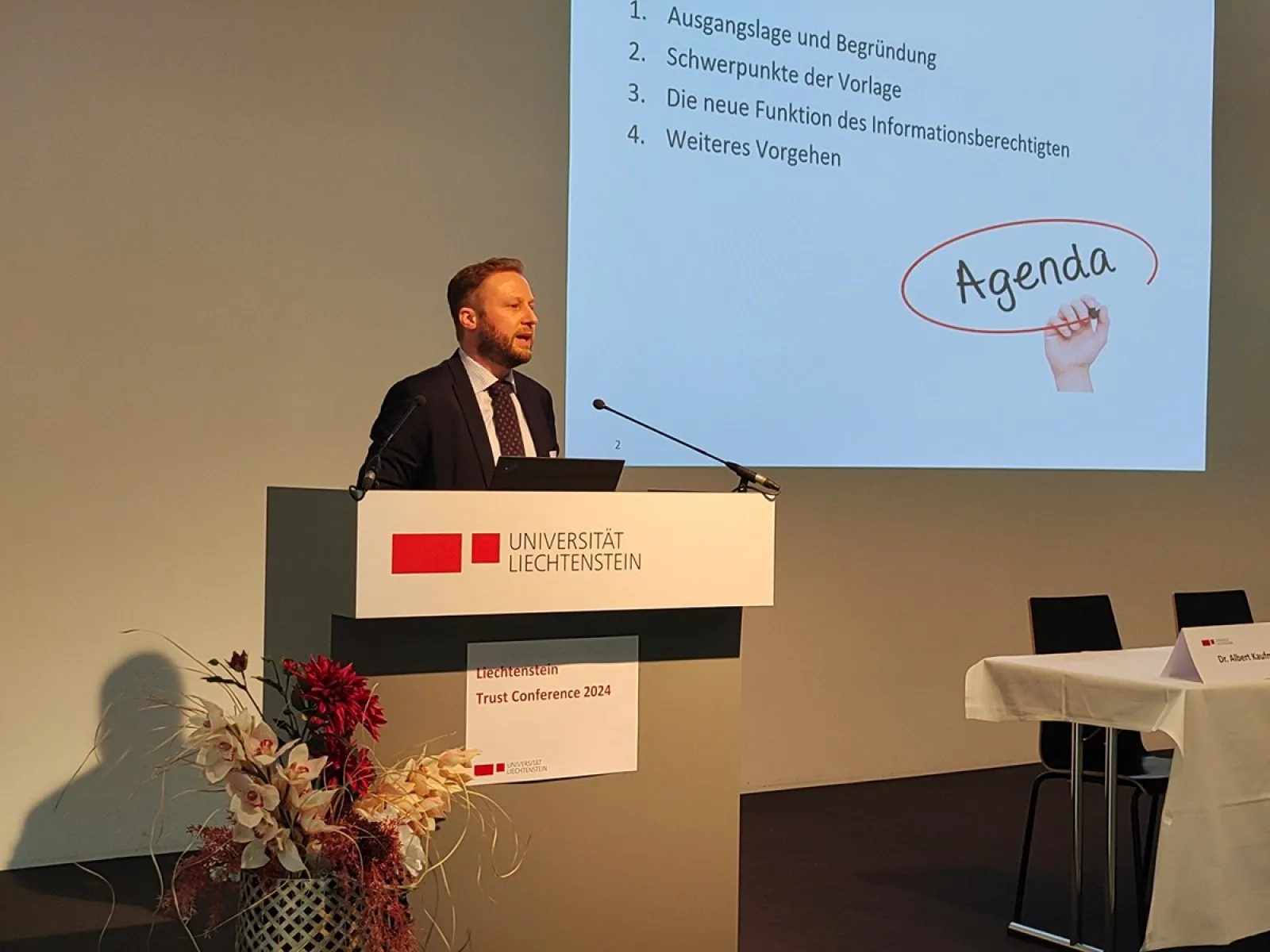 Ein Redner steht an einem Pult mit dem Logo der Universität Liechtenstein. Im Hintergrund ist eine Präsentationsfolie mit der Überschrift "Agenda" zu sehen. Links neben dem Pult steht eine Blumendekoration.