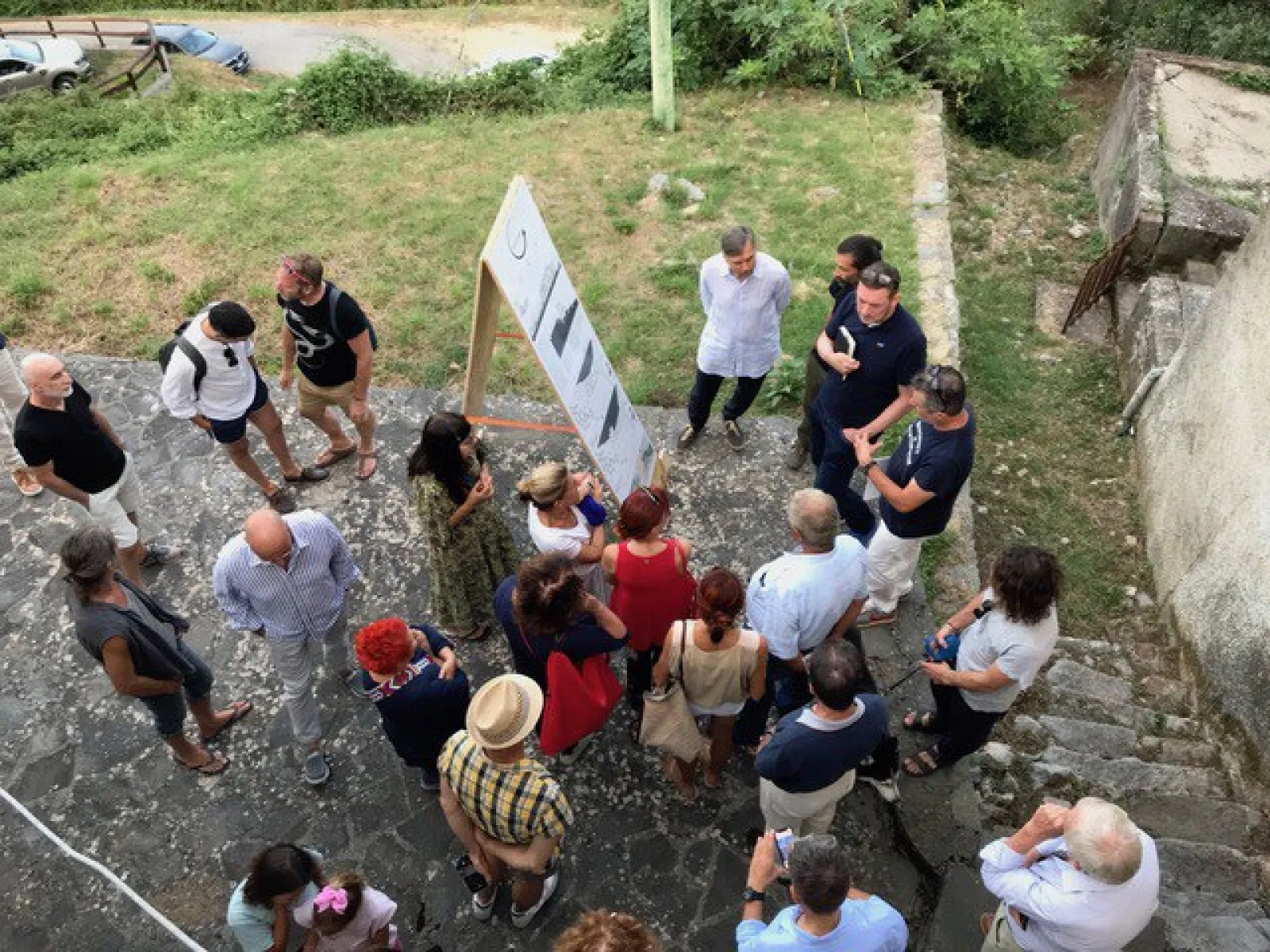 Eine Gruppe von Menschen steht im Freien um eine Tafel mit Bildern oder Plänen. Die Szene findet auf einem gepflasterten Platz mit angrenzendem Grünbereich statt. Die Personen scheinen in eine Diskussion oder Präsentation vertieft zu sein.