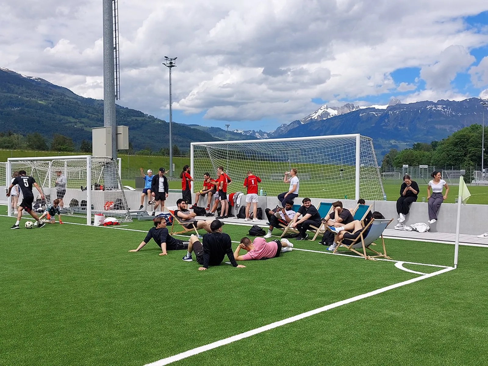 Teilnehmende des Völkerballturniers entspannen sich auf einem Sportplatz vor einer beeindruckenden Bergkulisse. Einige sitzen auf Liegestühlen, während andere am Spielfeldrand stehen. Im Hintergrund sind Fußballtore und Spieler in Sportkleidung zu sehen.