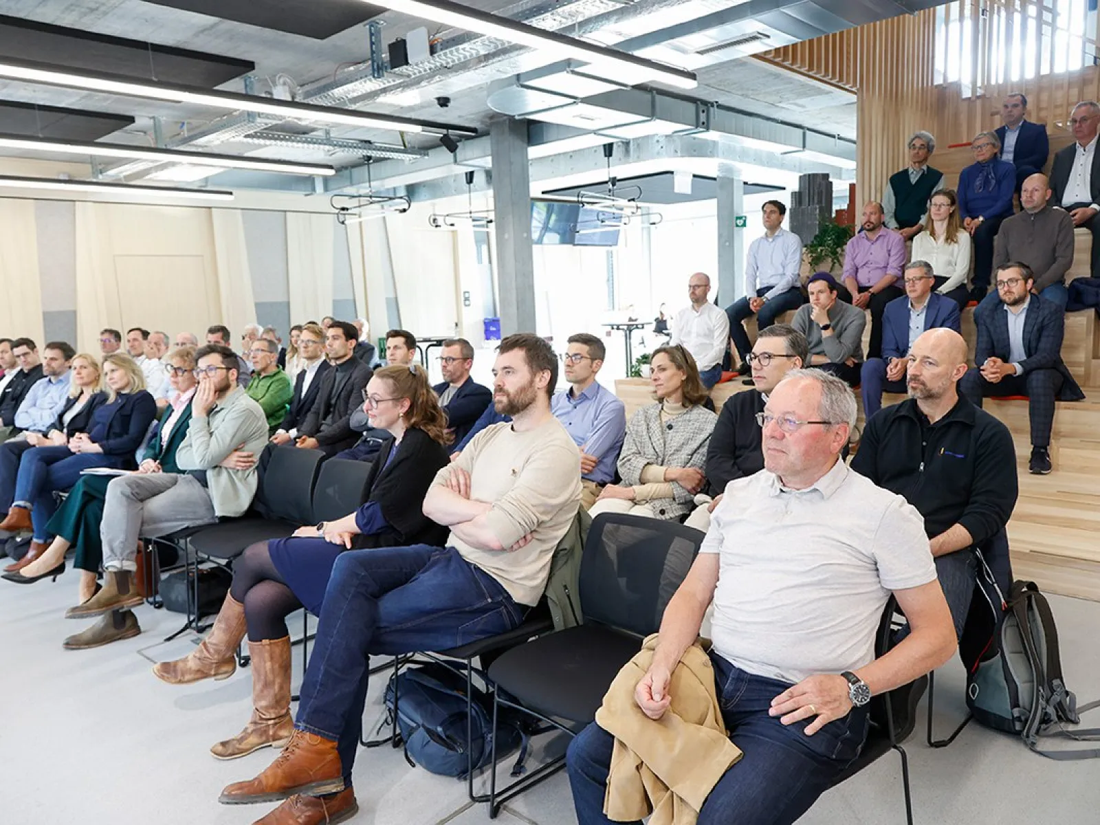 Eine Gruppe von etwa 50 Personen sitzt in einem modernen Konferenzraum und hört aufmerksam einer Präsentation zu. Die Veranstaltung findet an der Universität Liechtenstein statt und thematisiert den Wohnungsmarkt. Im Hintergrund sind einige Personen auf einer Treppe sitzend zu sehen.
