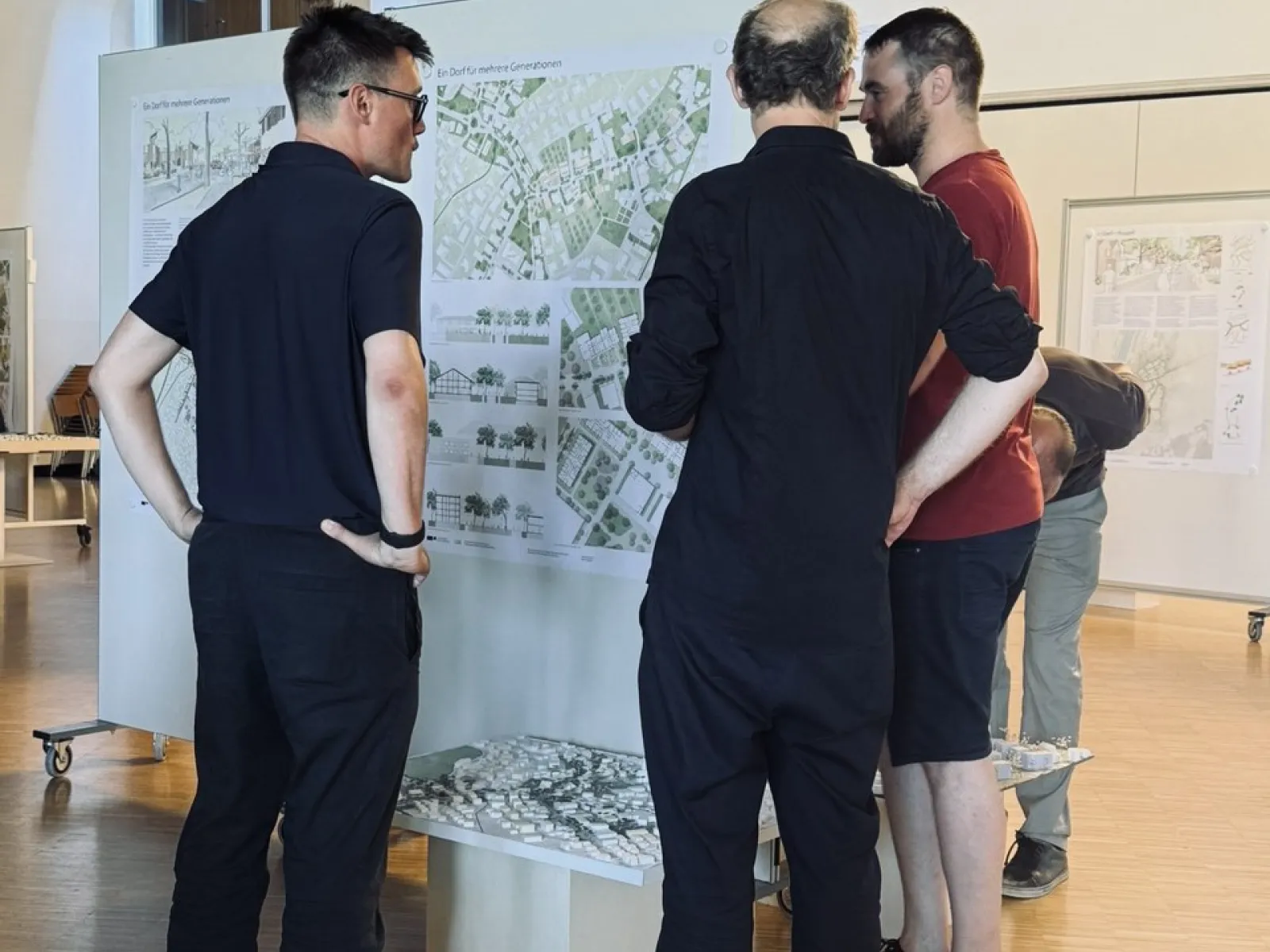 Drei Personen betrachten Architekturpläne und ein Modell in einem Ausstellungsraum der Universität Liechtenstein. Die Szene zeigt eine Diskussion über ortsbauliche Strategien zur Entwicklung des Zentrums von Ruggell.