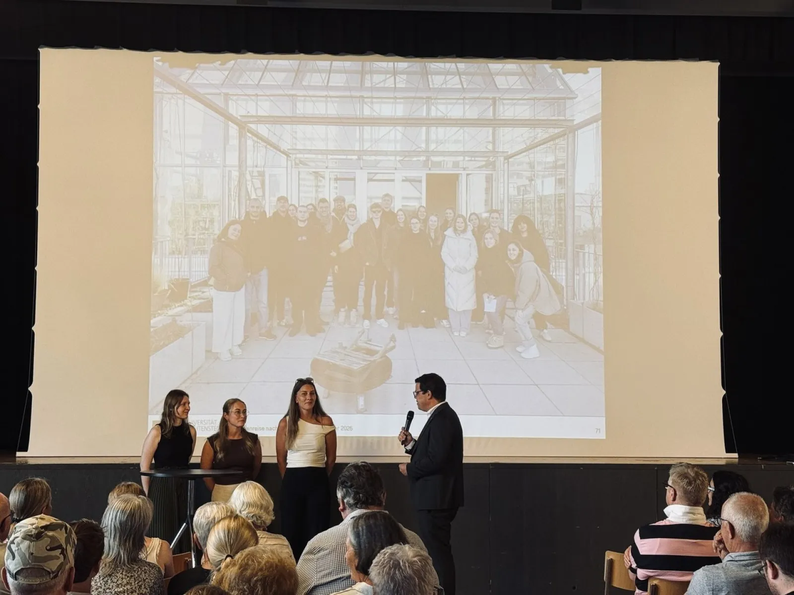 Eine Gruppe von Studierenden präsentiert ihre Projekte zur Ortsentwicklung in Ruggell vor einem Publikum. Im Hintergrund ist ein Bild der Studierenden auf einer Leinwand zu sehen.
