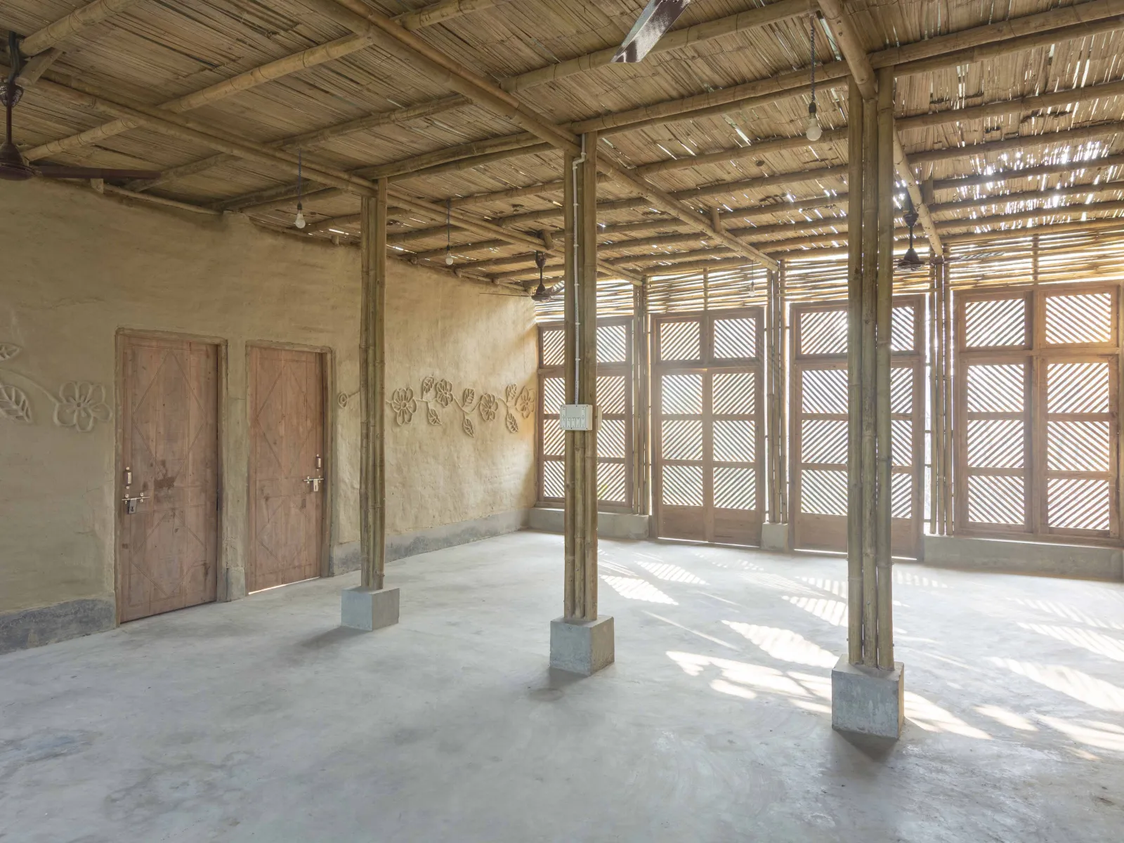 Ein sonnendurchfluteter Raum aus Bambus und Lehm mit zwei Holztüren und dekorativen Blumenreliefs an der Wand.