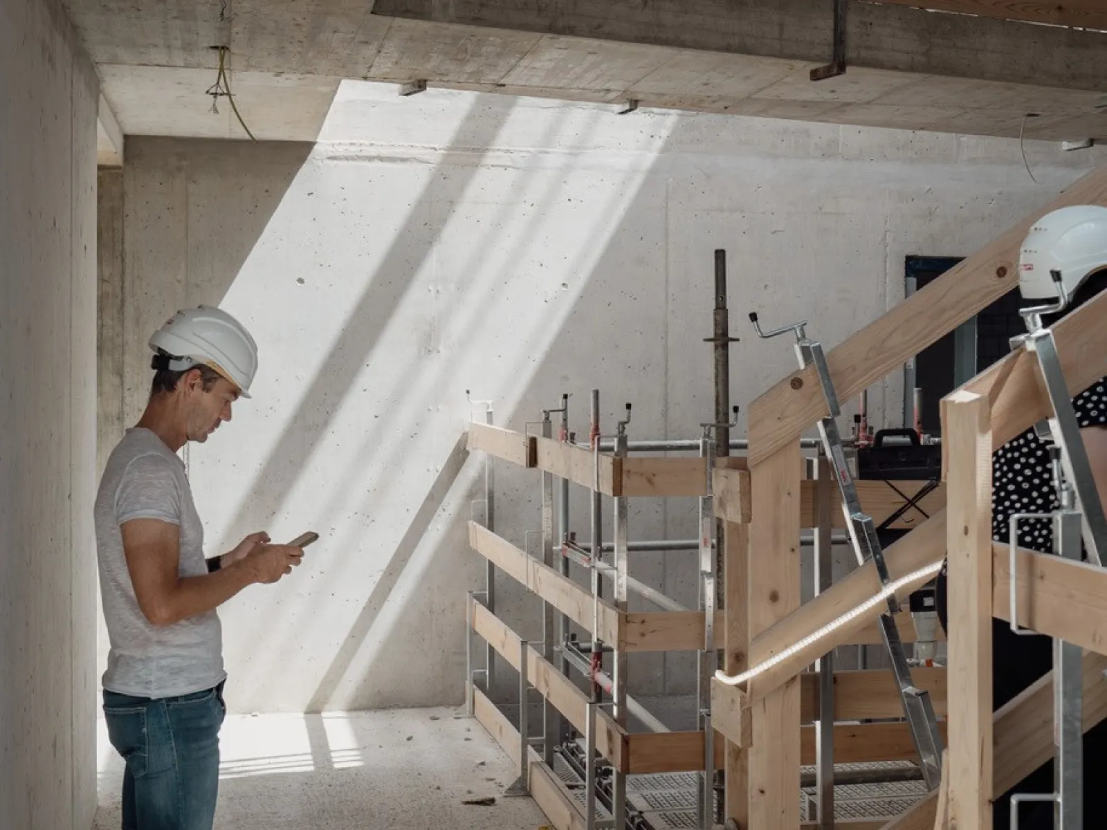 Ein Mann mit Bauhelm steht auf einer Baustelle und schaut in sein Handy 