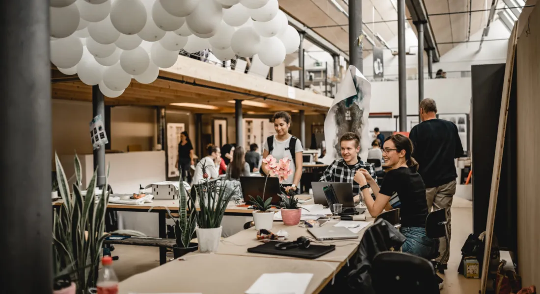 Eine Gruppe von Studierenden arbeitet gemeinsam in einem hellen, offenen Architekturstudio, das von natürlichem Licht durchflutet ist, mit weißen Luftballons, die von der Decke hängen.