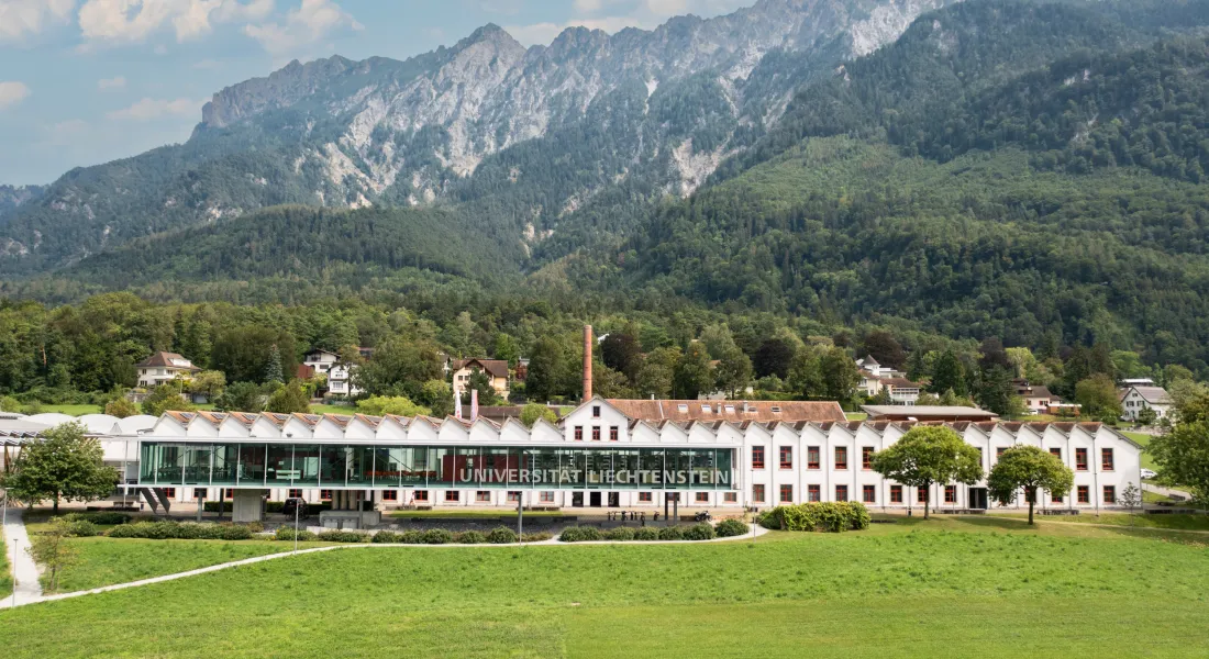Frontansicht der Universität Liechtenstein mit markanter Glasfassade und grüner Wiese im Vordergrund.