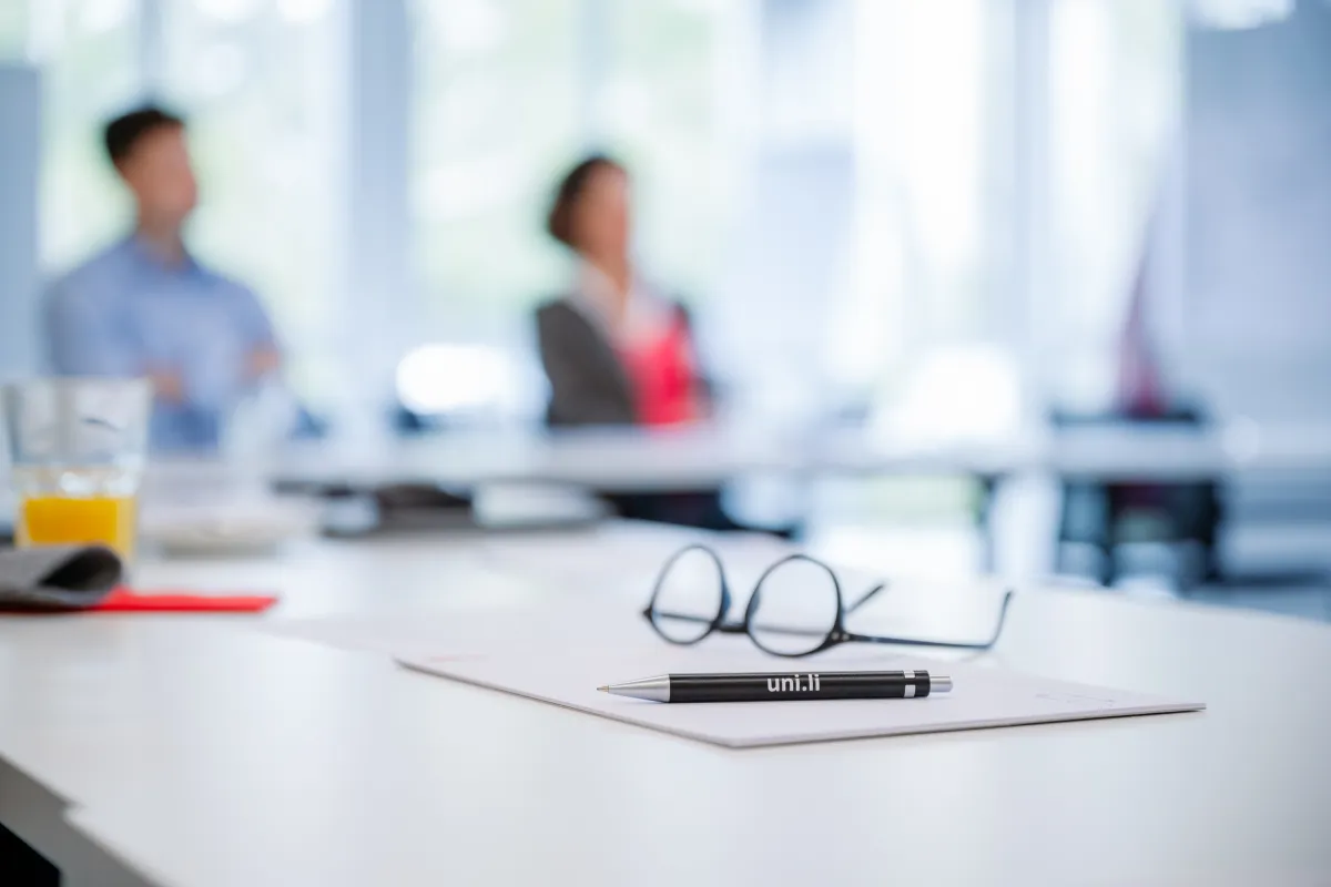 Brille und Stift mit der Aufschrift „uni.li“ auf einem Tisch in einem Seminarraum