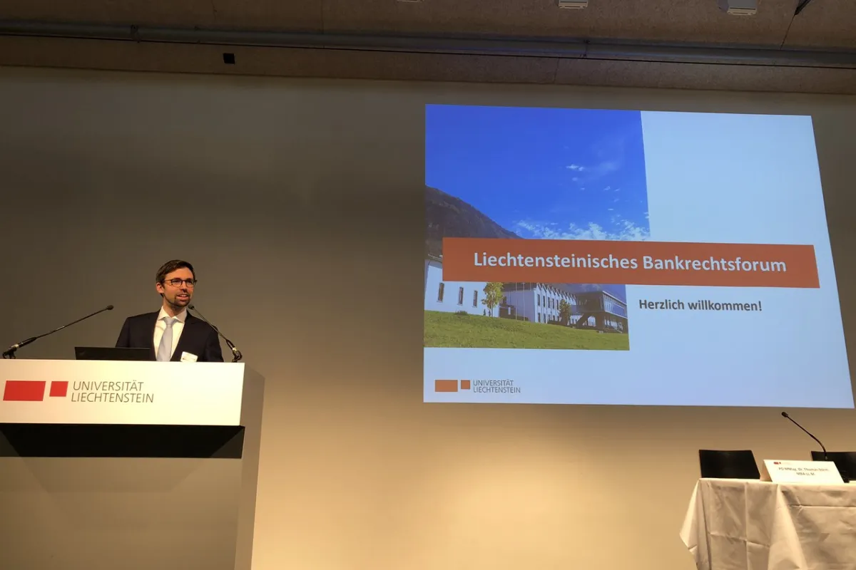 Ein Redner steht am Podium der Universität Liechtenstein während des Liechtensteinischen Bankrechtsforums. Auf der Leinwand ist eine Präsentationsfolie mit der Aufschrift „Liechtensteinisches Bankrechtsforum – Herzlich willkommen!“ zu sehen.