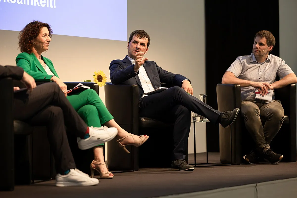 Podiumsdiskussion an der Universität Liechtenstein: Drei Personen sitzen auf einer Bühne in schwarzen Sesseln und diskutieren angeregt. Eine Frau in grünem Anzug und zwei Männer in formeller Kleidung sind zu sehen. Im Hintergrund ist ein Teil einer Leinwand mit Text sichtbar.